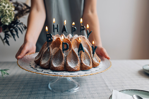 An anonymous white woman holding a cake stand with a bundt cake with lit birthday candles on it.