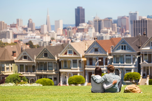 San Francisco tourist attraction at Alamo Square, the Painted Ladies famous postcard row, California travel. Couple tourists relaxing in grass enjoying popular destination. People lifestyle
