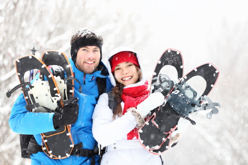 Winter couple happy outdoor hiking in snow on snowshoes. Healthy lifestyle photo of young smiling active mixed race couple snowshoeing outdoors. Asian woman, caucasian man.