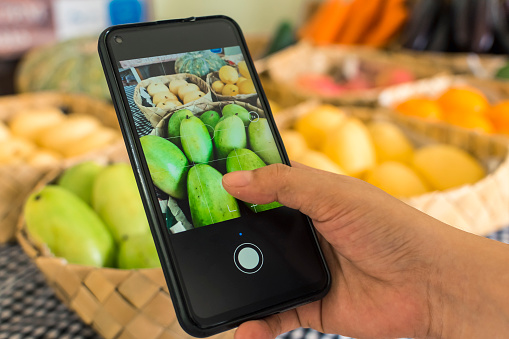 A man takes a picture of green mangoes and other produce with a cellphone at a fruit stand at a market.