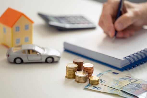 A MALE HAND WRITING IN THE NOTEBOOK NEXT TO A CALCULATOR, A LITTLE HOUSE, A TOY CAR AND SEVERAL STACKS OF EURO COINS AND NOTES. HOME ECONOMY CONTROL CONCEPT. FOCUS SELECTED. A MALE HAND WRITING IN THE NOTEBOOK NEXT TO A CALCULATOR, A LITTLE HOUSE, A TOY CAR AND SEVERAL STACKS OF EURO COINS AND NOTES. HOME ECONOMY CONTROL CONCEPT. FOCUS SELECTED. invoice pad stock pictures, royalty-free photos & images