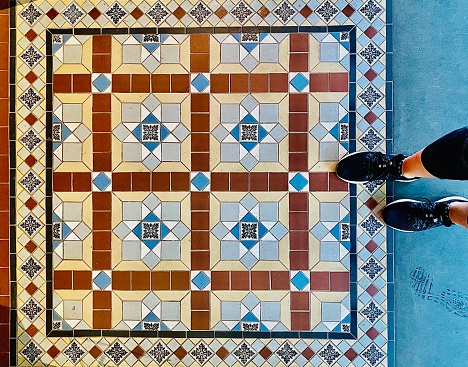 Horizontal flat lay looking down to square building entrance design of ceramic mosaic patterned tiles in blue and terracotta red square geometric shape with woman’s lower legs and feet in black trainers