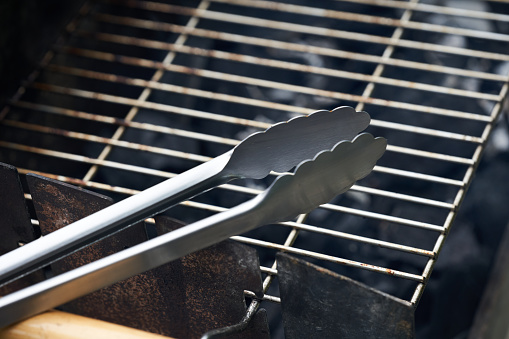 Stainless steel grill tongs on a dirty grill rust. Wooden handle blurred in the foreground.