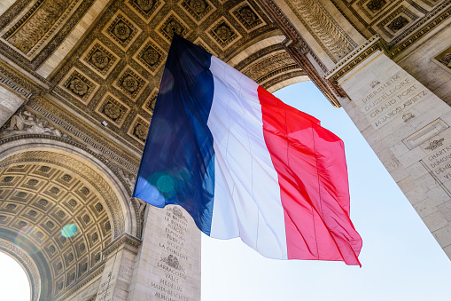 Low angle view of a large french flag fluttering in the wind under the vault of the Arc de Triomphe in Paris, France.