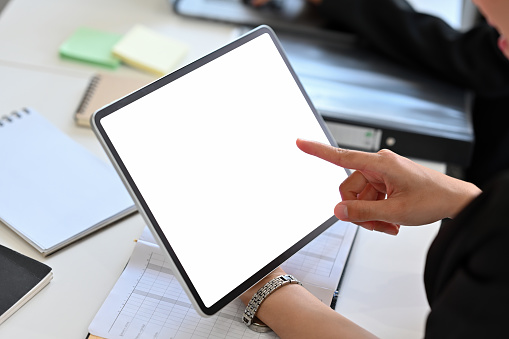 Cropped image of a woman holding a white blank screen digital tablet at the cluttered working desk.