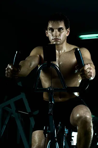 beautifull strong athletic guy,  execute exercise on exercise-bicycle, in  sport-hall, dapk background, hard light