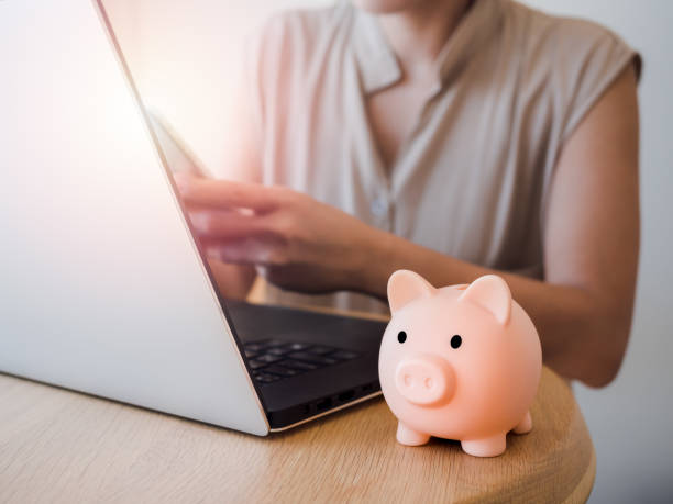 istock Pink piggy bank on wooden desk with woman's hands using a smart mobile phone while using a laptop computer on Investment technology, money transfer, savings, donate, online shopping concepts. 1373114764