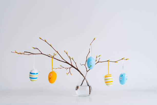 istock Minimalistic Easter still life. A glass vase with a branch on which colorful Easter eggs hang on a white background. Interior photography. 1373125643