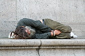 A bearded homeless man laying down on a stone ledge