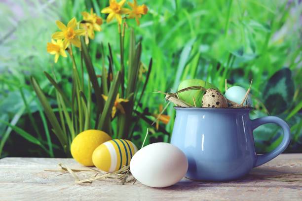 istock Easter eggs, a cup with Easter decorations and a flower, on a wooden table, against the background of green grass and blooming yellow daffodils 1373136074