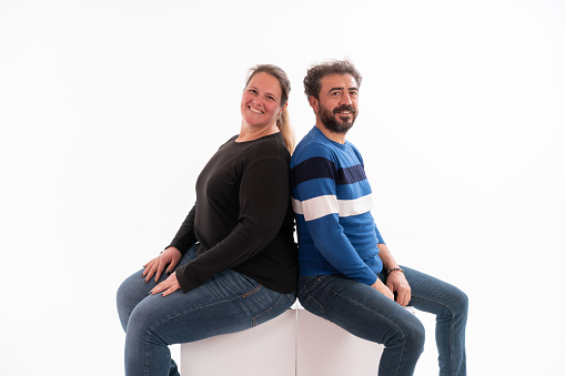 Studio shot of happy couple sitting on cube together at isolated white background.