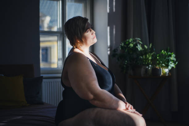 mujer gorda solitaria y deprimida sentada y mirando por la ventana de casa. - mature adult sadness overweight women fotografías e imágenes de stock