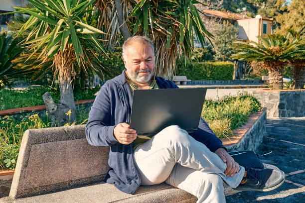 istock Handsome bearded mature man with laptop computer working outdoors while sitting on bench at the seaside. Modern lifestyle, connection, business, freelance work concept. 1373143302