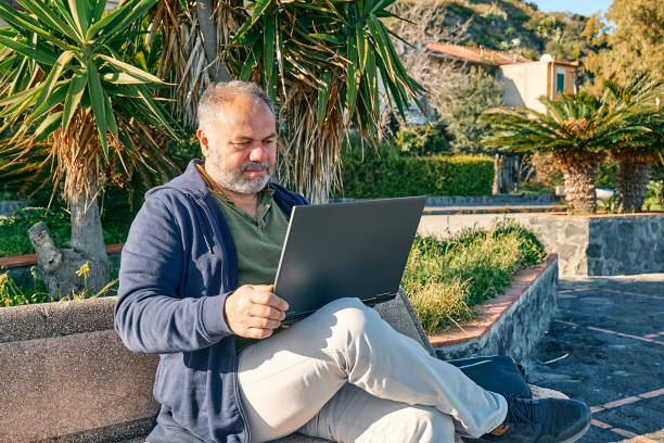 istock Handsome bearded mature man with laptop computer working outdoors while sitting on bench at the seaside. Modern lifestyle, connection, business, freelance work concept. 1373143375