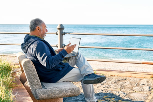 istock Bearded mature man with laptop working outdoors while sitting on bench at seaside.  Modern lifestyle, connection, business, freelance work concept. Smiling gray-head man having video conference 1373143426