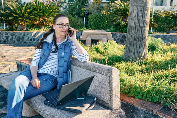 istock Sporty woman wearing eyeglasses with laptop computer working outdoors, talking on phone while sitting on bench at the seaside. Modern lifestyle, connection, blogging, business, freelance concept. 1373143602