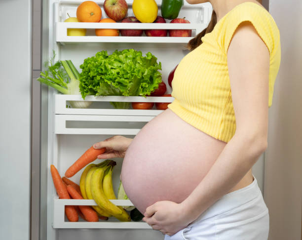Pregnant woman takes vegetables from the fridge, hungry woman takes carrot from the fridge full of vegetables and fresh fruit, healthy diet in maternity. stock photo
