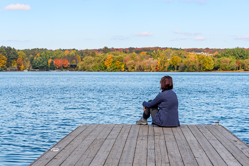 Woman sitting on a wooden pier on a lake on a clear autumn day. Concept of loneliness. Huntsville, On, Canada.