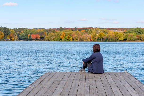 Lonely woman sitting on a wooden jetty on a sunny autumn day Woman sitting on a wooden pier on a lake on a clear autumn day. Concept of loneliness. Huntsville, On, Canada. huntsville ontario stock pictures, royalty-free photos & images