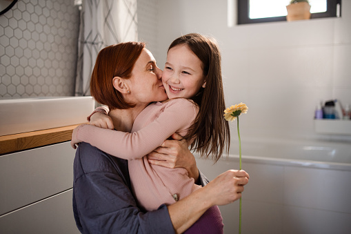 A little girl congratulates mother and gives her flower in bathroom at home.