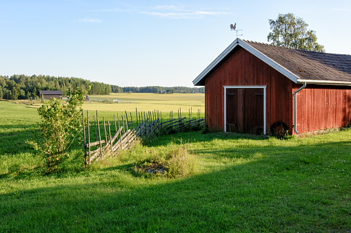 Traditional Finnish red wooden barn with roundpole fence and the weather vane on the roof. Old idyllic farm in the countryside.