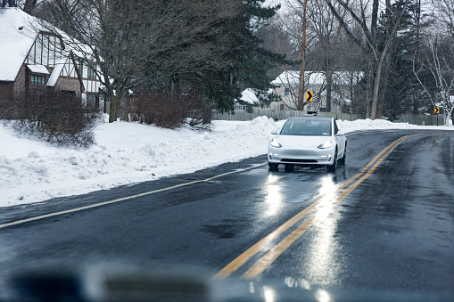 A white Tesla Model 3 sedan BEV (Battery Electric Vehicle - fully powered purely by electricity) with brightly illuminated headlights reflecting across the slippery wet asphalt is approaching on a rural road during a sloppy, drizzly winter snow storm in western New York State. Photo taken on Clark Road in Penfield, New York near Rochester, NY about 4:45pm on February 18, 2021.
