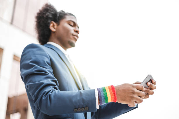 jeune homme d’affaires africain avec bracelet lgbt utilisant un smartphone à l’extérieur. concept de diversité. - homosexual gay pride business rainbow photos et images de collection