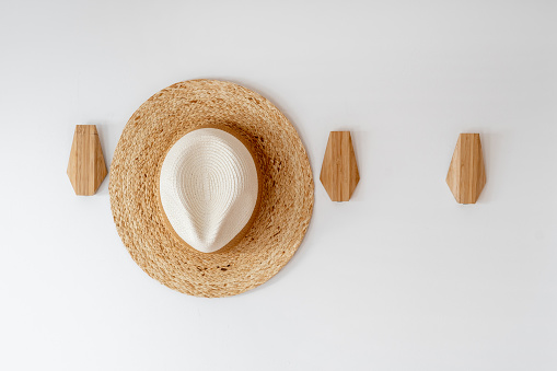 Sunhat on hanger for clothes in hall. Close up view of brimmed hat hanging on wooden hook for apparel on white copy space wall in room