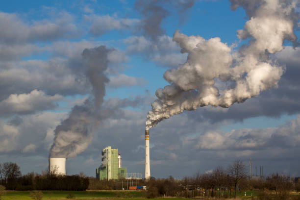 Aerial drone view of a large coal-fired power plant with multiple cooling towers and tall smokestacks releasing steam and emissions beneath a dark, overcast sky. The industrial complex conveys a heavy, atmospheric mood, symbolizing fossil fuel energy production, carbon emissions, environmental impact, and climate change.