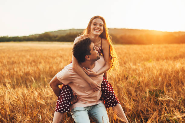 Caucasian couple man giving woman piggy back ride through the wheat field, together at sunset. Activity relationship. stock photo