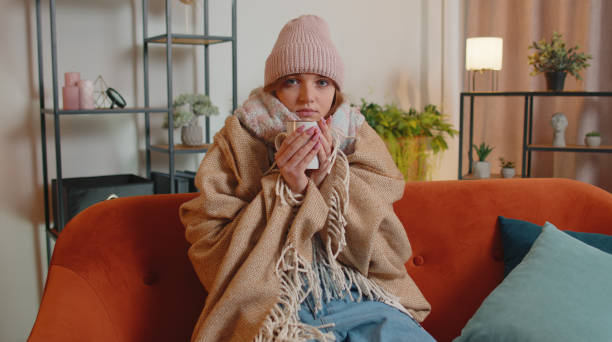 młoda chora kobieta nosi kapelusz owinięty w kratę siedzieć samotnie drżąc z zimna na kanapie pijąc gorącą herbatę - shivering zdjęcia i obrazy z banku zdjęć