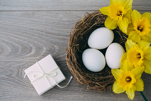 Copy space shot of a wooden table with a small arrangement on it. A little gift box with a nest with three white eggs and yellow daffodils as a decoration on it.