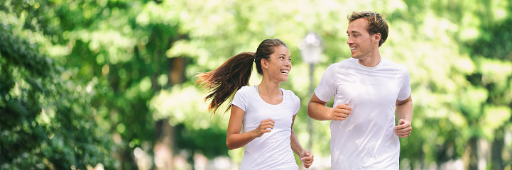 Run exercise fitness friends walking running talking together on fun race in city park panoramic banner background. Healthy active lifestyle young people, Asian woman, Caucasian man couple.