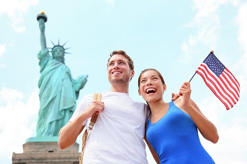 Tourist travel couple at Statue of Liberty, New York City, USA. Multiracial tourist couple on summer vacation holidays. Asian woman holding American flag smiling happy.