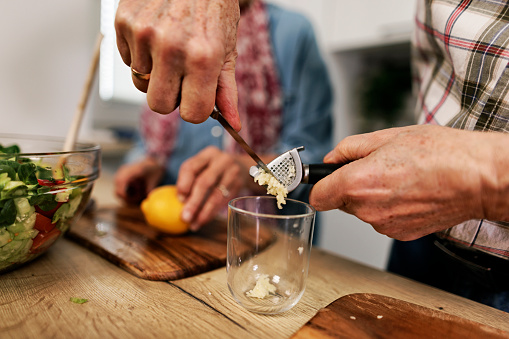 Senior couple is preparing a healthy vegetable salad at home. They are preparing the vinaigrette dressing. The man is pressing garlic to a glass.\nCanon R5