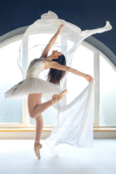 une jeune ballerine motivée vêtue d’un costume de tutu blanc pratique des mouvements de ballet au studio de ballet devant un grand fond de fenêtre en forme de rond dans une atmosphère naturelle de lumière du jour ambiante - round toe shoes photos et images de collection