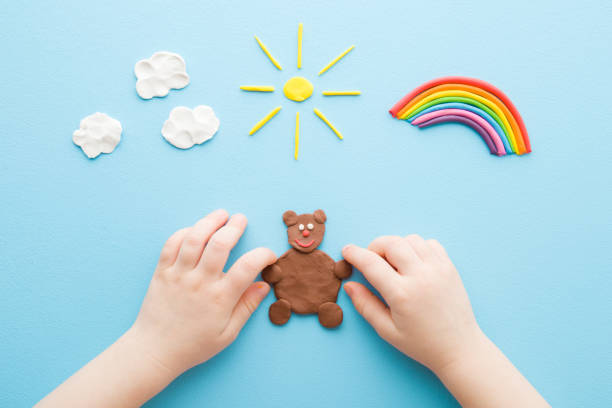 istock Baby hands making brown bear from modeling clay. Colorful rainbow, sun and white clouds on light blue table background. Pastel color. Closeup. Point of view shot. Toddler development. Top down view. 1373247780