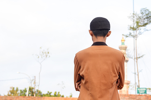 Back view of middle eastern man in brown muslim shirt and black cap looking to the far mosque with hand crossed