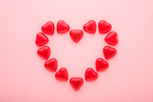istock Heart shape created from chocolate candies wrapped in bright red foil paper on light pink table background. Pastel color. Love concept. Closeup. Top down view. 1373251452