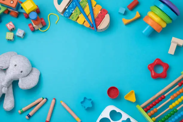 Top view of a large group of colorful toddler toys disposed at the borders of the image leaving a useful copy space at the center on a bluish background. The composition includes a xylophone, an abacus, a wooden train, a stuffed elephant,  and some crayons a some wooden letter blocks.
