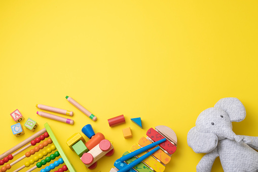 Top view of a group of colorful toddler toys disposed at the bottom of the image leaving a useful copy space at the top on a yellow background. The composition includes a stuffed elephant, a wooden abacus, a xylophone, some wooden crayons, a wooden train, and some wooden letter blocks.