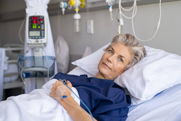 Senior woman resting on hospital bed Mature woman recovering from illness while lying on hospital bed with iv drip in hand. Portrait of old hospitalized patient with grey hair recovering after surgery. Happy senior woman lying on bed in hospital ward while looking at camera. sick woman hospital bed stock pictures, royalty-free photos & images