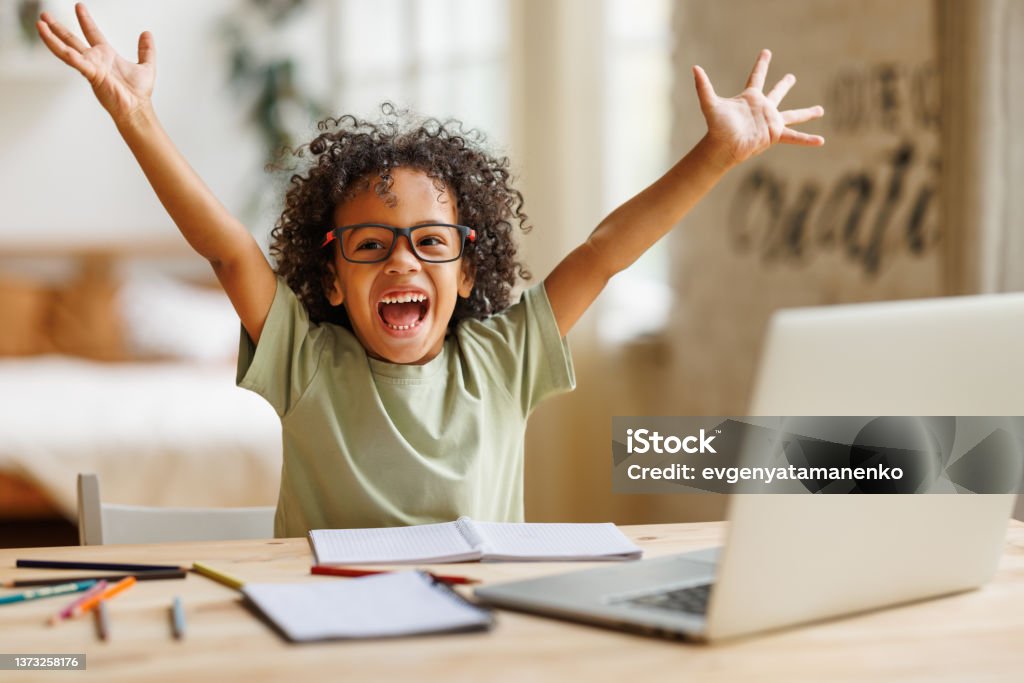Little african american school boy raising hands up with excitement during home distance education Happy excited little african american elementary school boy raising hands up and screaming with excitement during home distance education on laptop computer, kid celebrating finishing homework Child Stock Photo