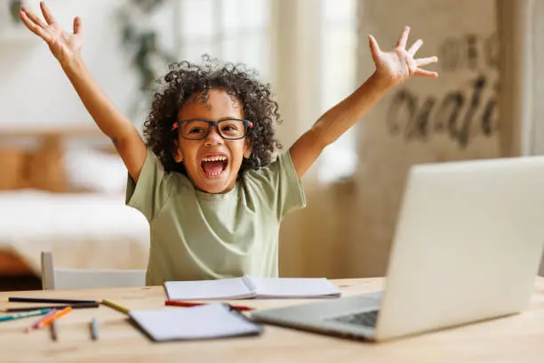 Photo of Little african american school boy raising hands up with excitement during home distance education