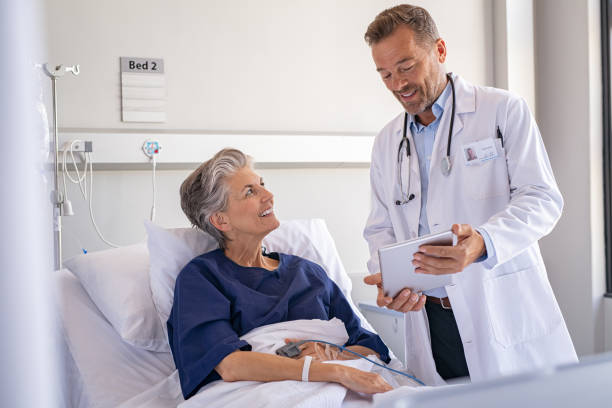 Doctor tells good news to patient Smiling senior woman sitting on hospital bed listening to mature surgeon at private clinic. Confident specialist in labcoat holding digital tablet and talking to old patient. Happy professional friendly doctor meeting elderly patient on a hospital bed and explaining health results. old woman smiling tablet stock pictures, royalty-free photos & images