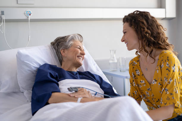 Woman visiting senior mother in hospital Young daughter visiting sick old mother in hospital. Happy senior patient smiling while in conversation with her lovely daughter. Granddaughter visiting and cheering her hospitalized grandmother lying in bed at hospital ward after surgery. sick woman hospital bed stock pictures, royalty-free photos & images