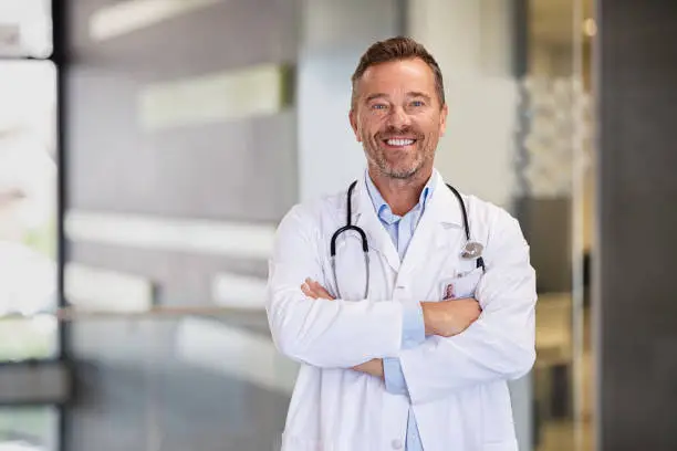 Portrait of happy mature doctor with folded arms standing at hospital hallway. Confident male doctor in a labcoat and stethoscope looking at camera with satisfaction. Smiling confident general practitioner at private health clinic.