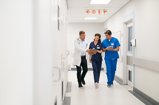 Medical team walking in hurry and interacting at modern hospital. Mature doctor and surgeon working on digital tablet with nurse and walking in hospital hallway. Head physician working with his medical team at clinic.