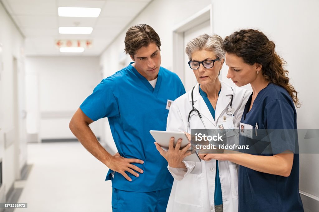 Doctor and nurse discussing patient case at hospital Mature female doctor discussing medical report with nurses in hospital hallway. Senior general practitioner discussing patient case status with group of medical staff after surgery. Doctor working on digital tablet while in conversation with healthcare workers, copy space. Doctor Stock Photo