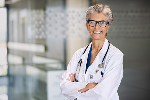 Portrait of proud female doctor with stethoscope around neck at medical clinic. Happy smiling senior woman doctor at hospital lobby looking at camera with copy space. Mature general practitioner or physician feeling confident.